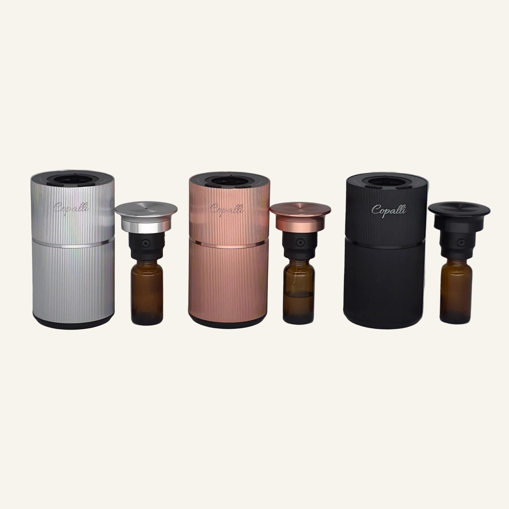 Three Copalli Mini diffusers in silver, rose gold, and black, shown open to reveal the internal fragrance oil bottles, set against a minimalist light cream background.