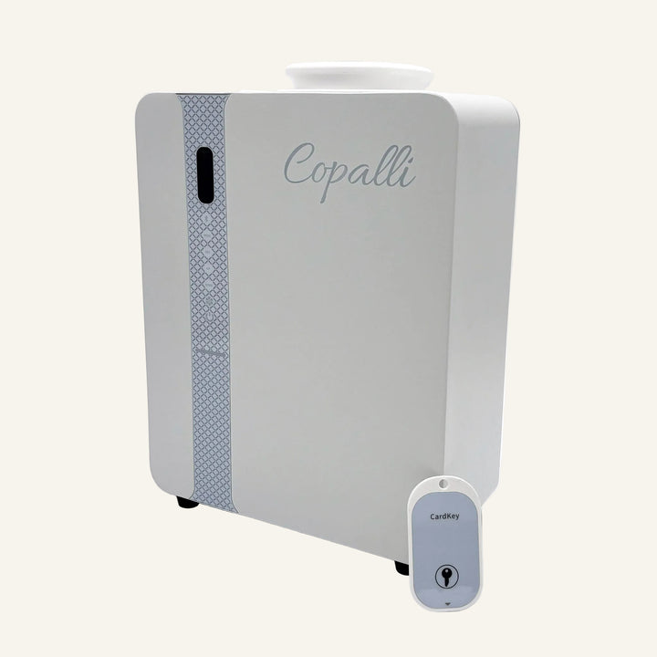 Our white Copalli Pro Plus Diffuser shown from a side profile, highlighting its industrial waterless design and minimalist branding, accompanied by its remote control on a light cream background.
