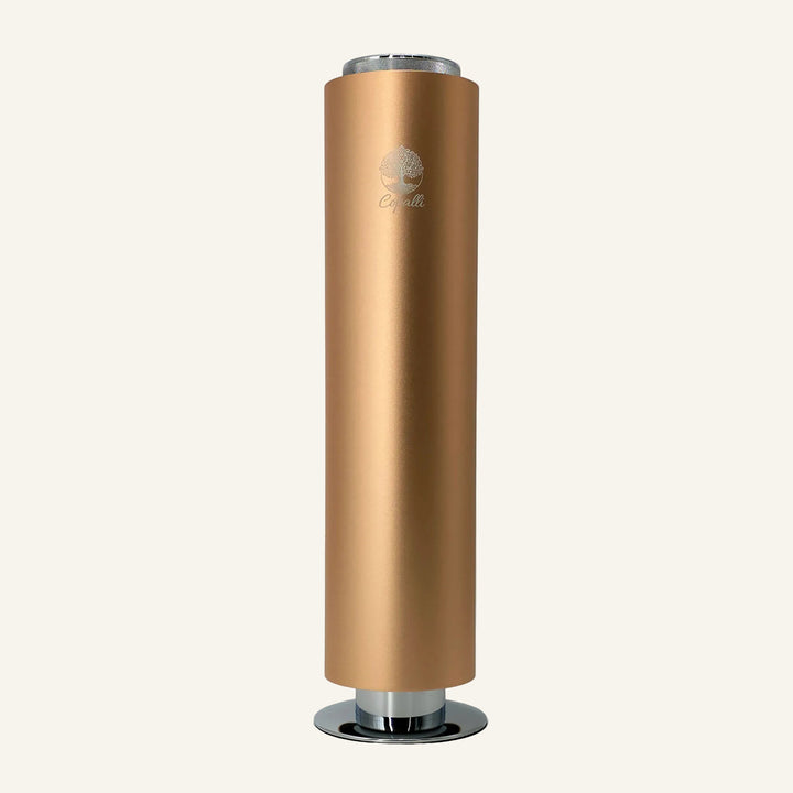 Our gold Tower Pro Diffuser by Copalli, featuring a tall cylindrical body with a gold metallic finish and a minimalist tree logo, centered on a light cream background.